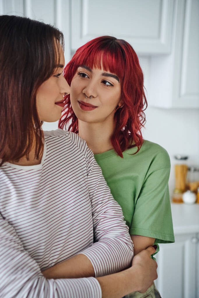 πορτρέτο της ευτυχισμένης και τρυπημένη λεσβία γυναίκα με κόκκινα μαλλιά κοιτάζοντας τη φίλη της στο σπίτι - Φωτογραφία, εικόνα