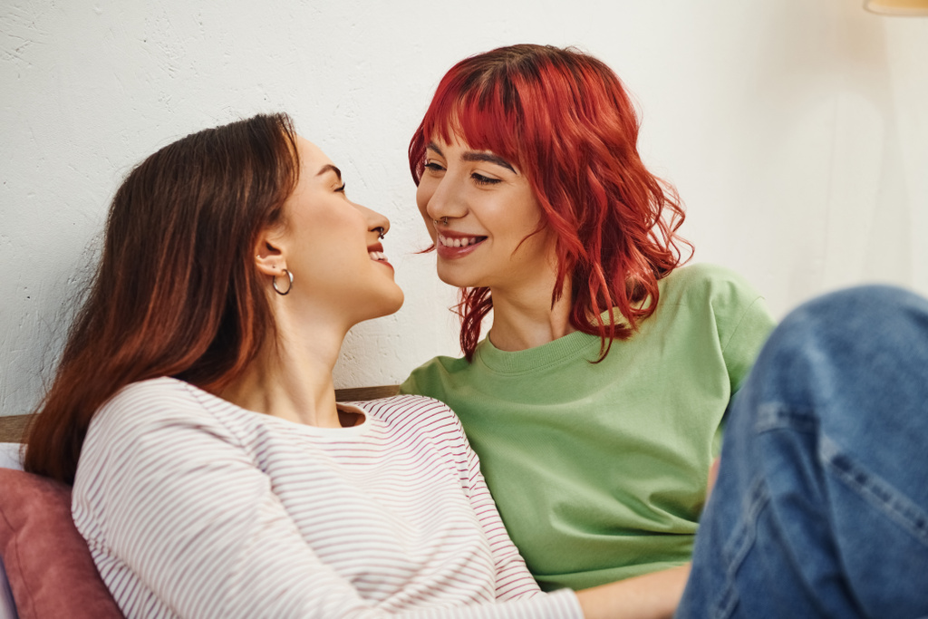alegre joven lesbiana pareja en casual desgaste mirando uno al otro mientras sentado juntos en la cama - Foto, Imagen