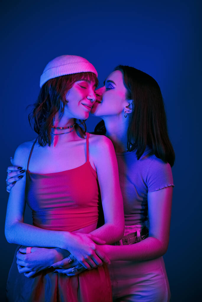 Mutlu lezbiyen çiftin mor ışıkları altında cesur makyajı, mutluluğu ve aşkı ile yanaklarında şehvetli bir öpücük. - Fotoğraf, Görsel