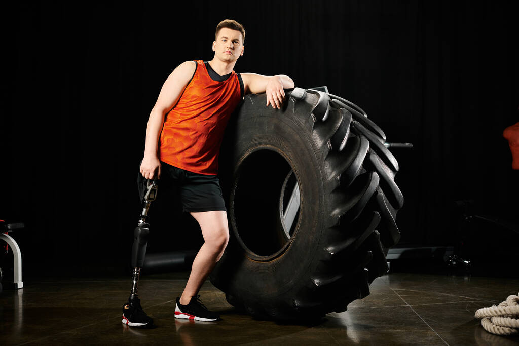 Człowiek z protezą nogi stoi pewnie obok kolosalnej opony na siłowni, pokazując swoją siłę i determinację.. - Zdjęcie, obraz