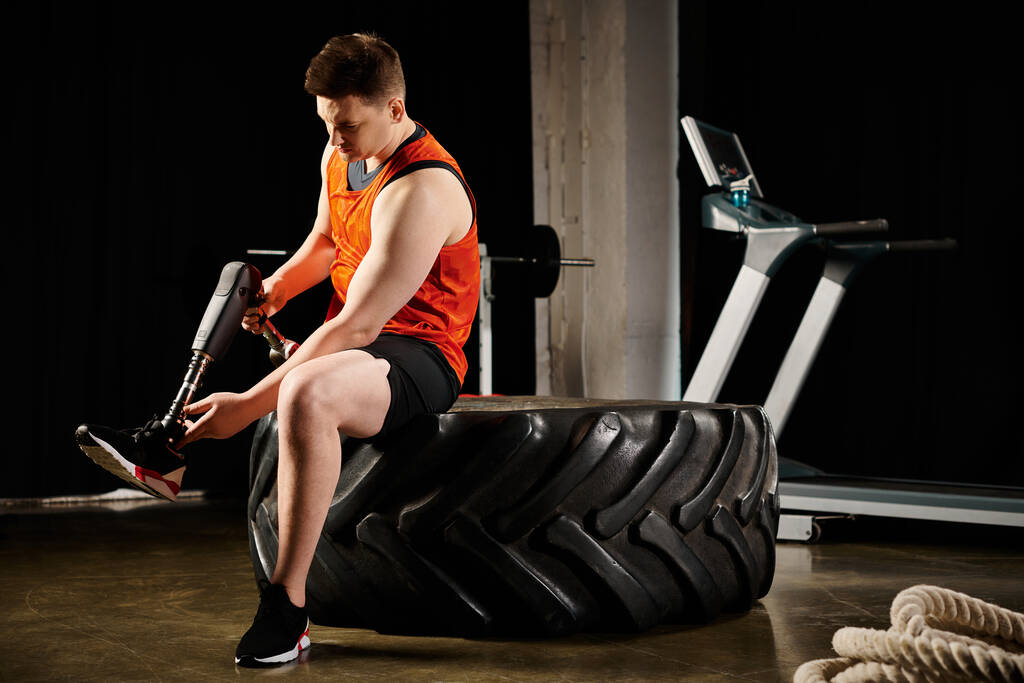 Un hombre discapacitado con una pierna protésica sentado con confianza en un neumático, haciendo ejercicio en el gimnasio. - Foto, imagen