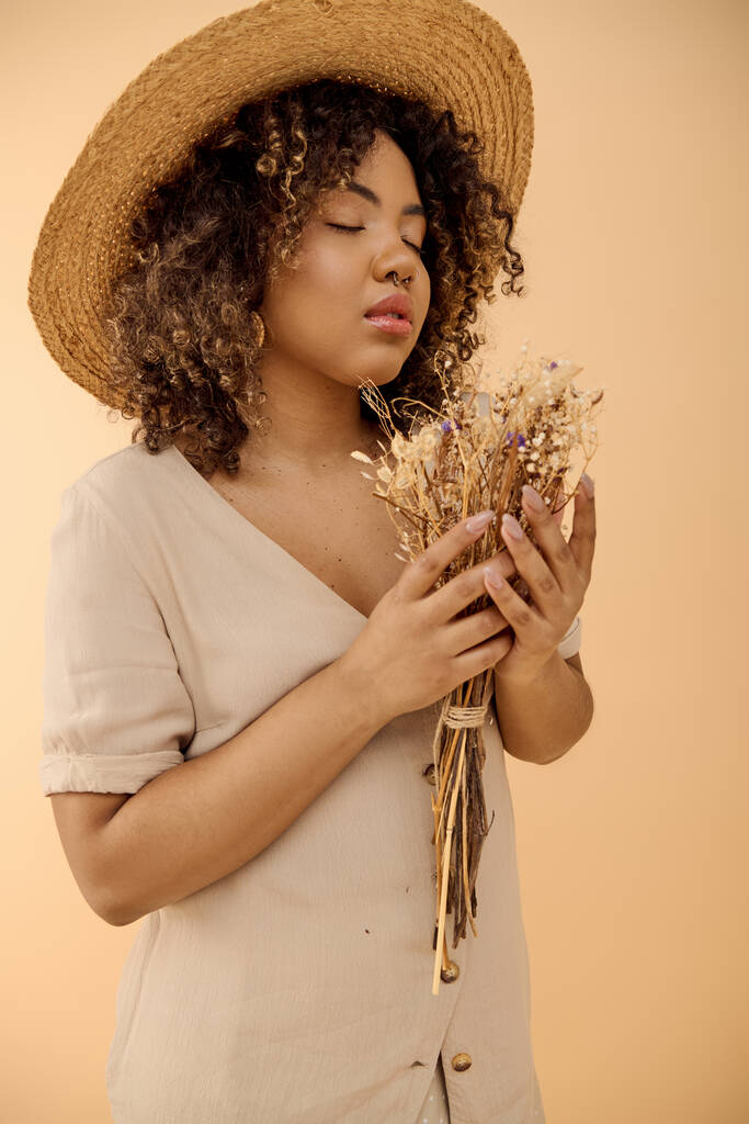 Eine hinreißende junge Afroamerikanerin mit lockigem Haar, trägt einen Strohhut und hält einen Strauß getrockneter Blumen in der Hand.. - Foto, Bild