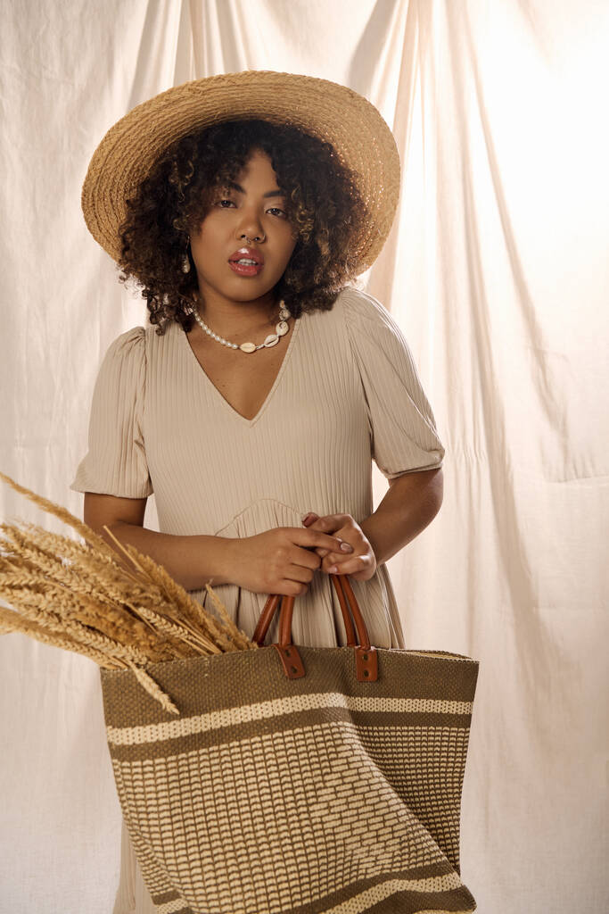 Una mujer afroamericana elegante con el pelo rizado, con un sombrero de paja y sosteniendo una bolsa en un entorno de estudio. - Foto, imagen