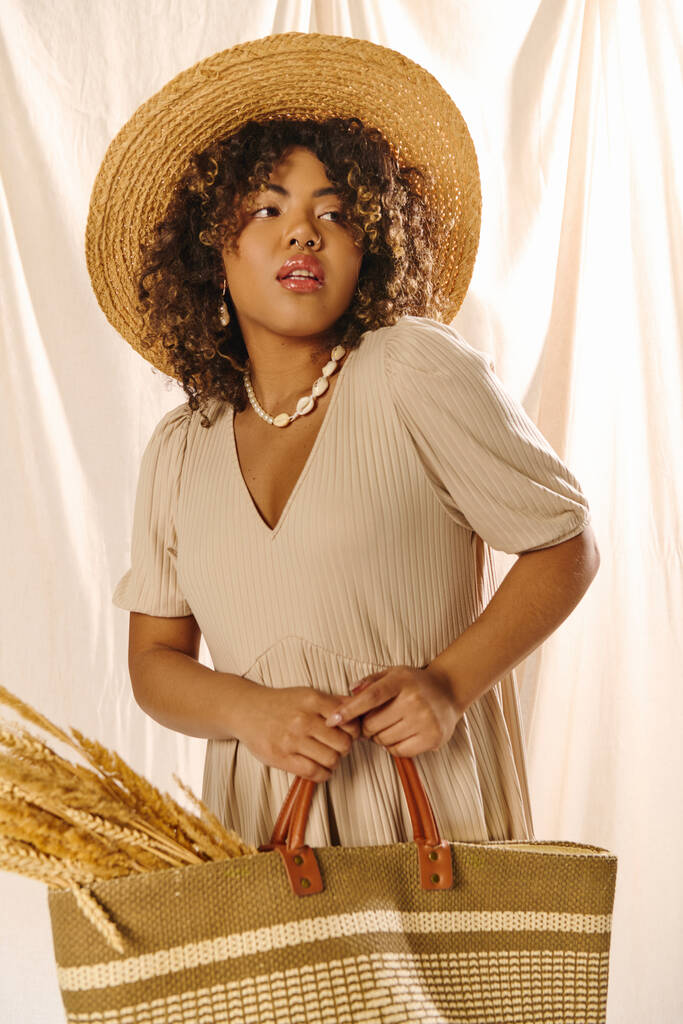 Una joven afroamericana con el pelo rizado posando elegantemente en un sombrero de paja mientras sostiene una bolsa de paja en un ambiente de estudio. - Foto, Imagen