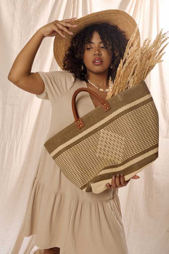 Όμορφη Αφροαμερικανή γυναίκα με σγουρά μαλλιά σε λευκό φόρεμα, κρατώντας με χάρη μια κομψή τσάντα άχυρο. - Φωτογραφία, εικόνα