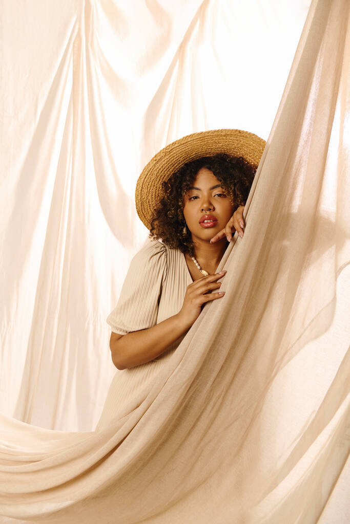 Eine schöne junge Afroamerikanerin mit lockigem Haar und Strohhut guckt aus einem Vorhang in einem Studio-Setting. - Foto, Bild