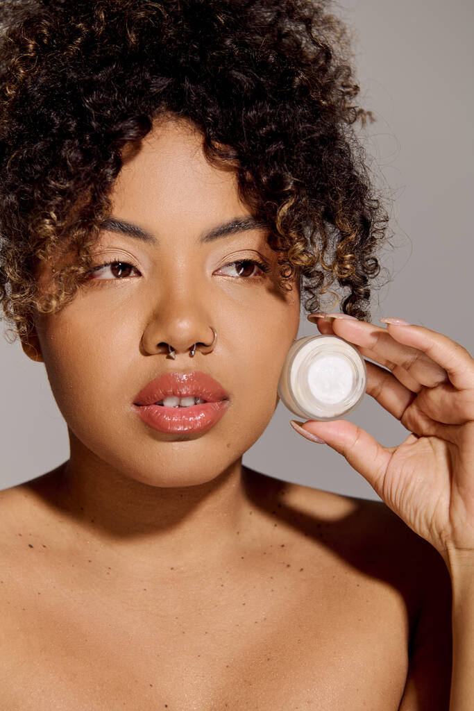 Una joven afroamericana con el pelo rizado sostiene un frasco de crema frente a su cara, haciendo hincapié en el cuidado de la piel y la belleza. - Foto, imagen