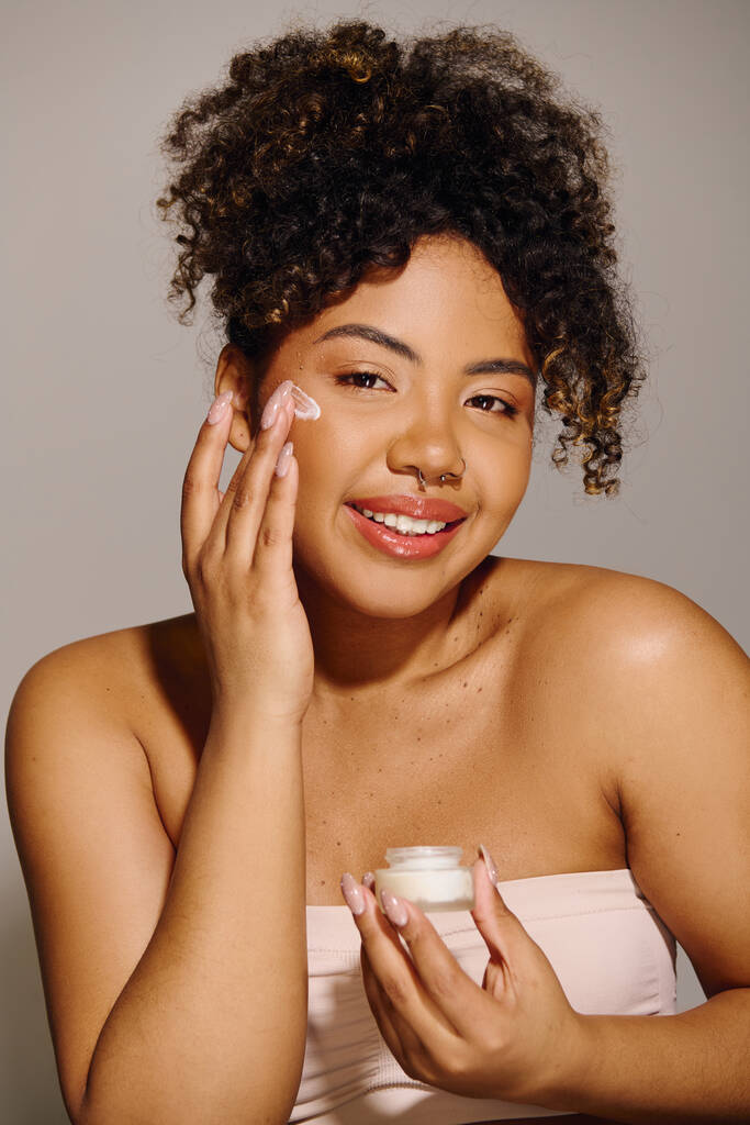 Beautiful African American woman with curly hair holding a jar of cream in front of her face in a studio setting. - Photo, Image