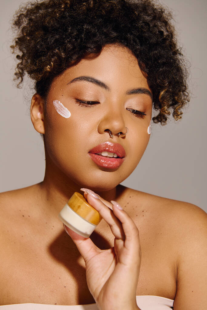 Jeune femme afro-américaine aux cheveux bouclés appliquant de la crème d'un bocal sur son visage dans un cadre de studio. - Photo, image