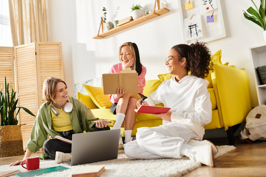 Wielokulturowe nastolatki siedzą razem na żółtej kanapie, studiując i dzieląc się wiedzą w przytulnym domu. - Zdjęcie, obraz