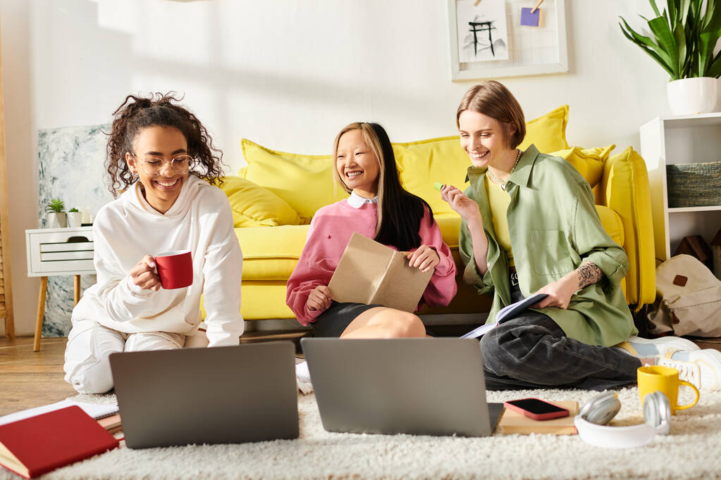 Τρία έφηβα κορίτσια διαφορετικών φυλών κάθονται στο πάτωμα με φορητούς υπολογιστές, ασχολούνται με τις σπουδές τους και την προώθηση ενός δεσμού φιλίας μέσω της εκπαίδευσης. - Φωτογραφία, εικόνα