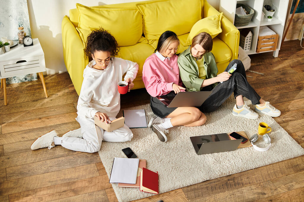 Tres adolescentes diversas se sientan en el suelo frente a un sofá amarillo, absortas en estudiar juntas. - Foto, Imagen