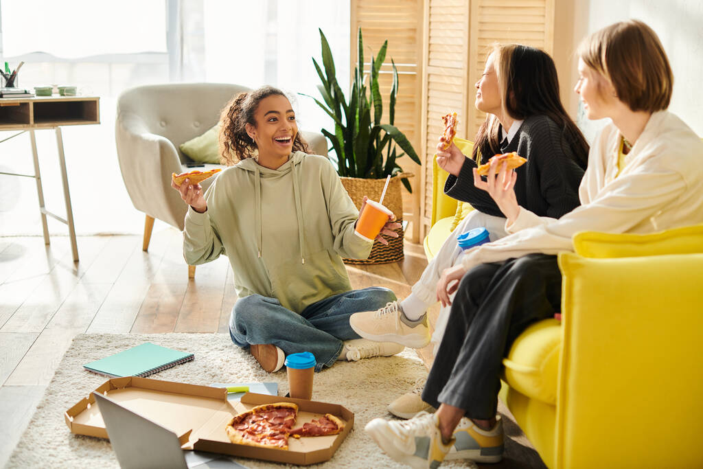 Çok kültürlü genç kızlar rahat bir oturma odasında toplanıp pizza dilimleriyle kaynaşıyor ve kahkahalarını paylaşıyorlar.. - Fotoğraf, Görsel