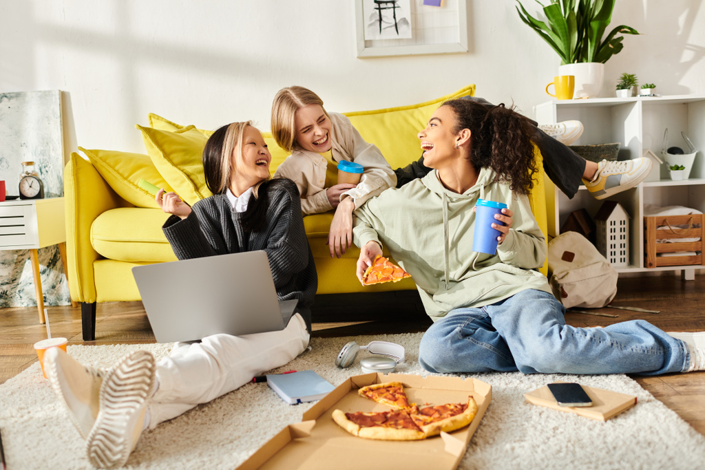 Zróżnicowana grupa nastolatek śmieje się i rozmawia na kanapie, pijąc pizzę i pijąc w przytulnym otoczeniu domowym. - Zdjęcie, obraz