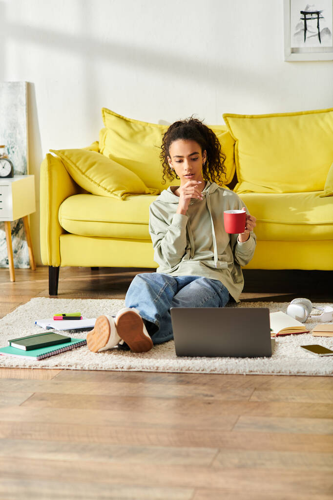 Młoda kobieta siedzi na podłodze, z wdziękiem trzymając filiżankę ciepłej kawy podczas nauki na laptopie w domu. - Zdjęcie, obraz