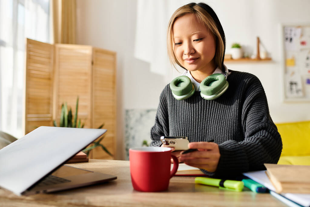 Egy fiatal ázsiai nő ül az asztalnál, egy csésze kávét kóstolgat, miközben otthon egy laptoppal tanul.. - Fotó, kép