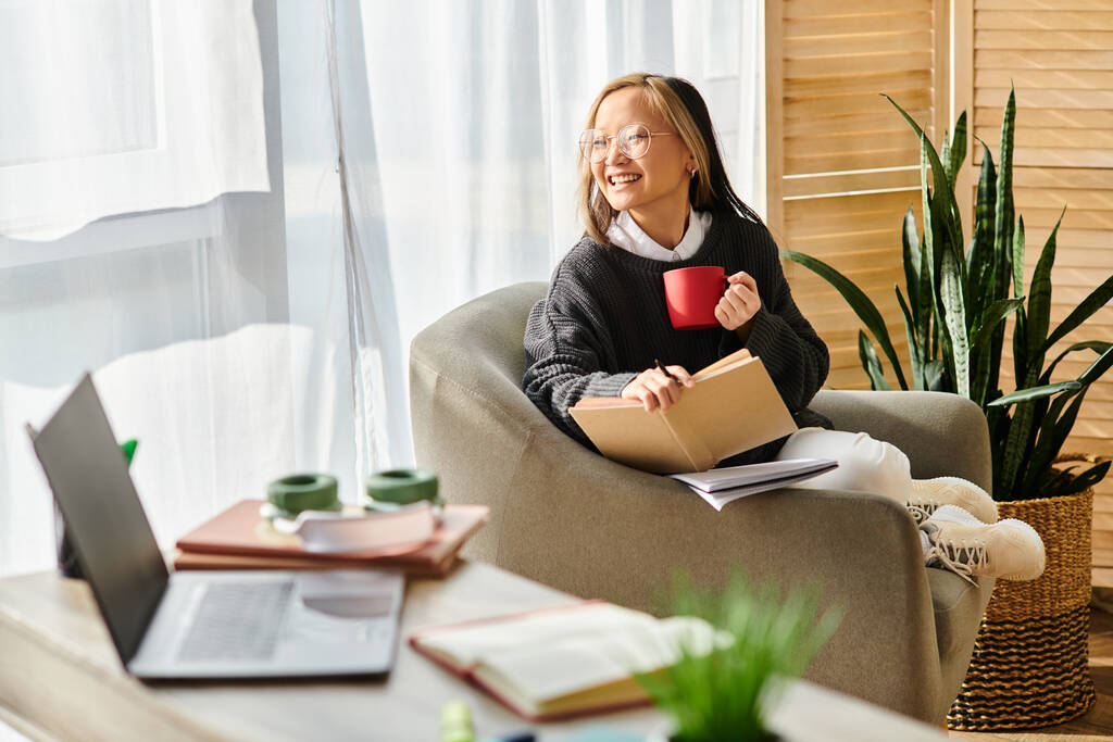 Egy fiatal ázsiai nő ül egy székben, könyv nyitva előtte, kezében egy gőzölgő csésze kávé. - Fotó, kép