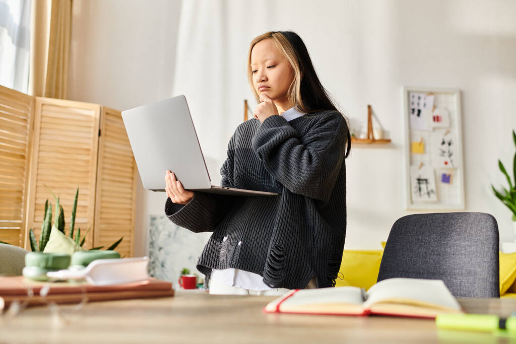 Молодая азиатка стоит перед ноутбуком, поглощенная электронным обучением дома. - Фото, изображение