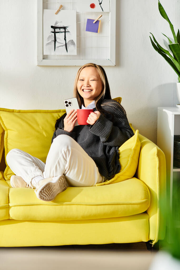 Nuori aasialainen nainen opiskelee kotona, istuu keltaisella sohvalla, pitäen kuppia. - Valokuva, kuva