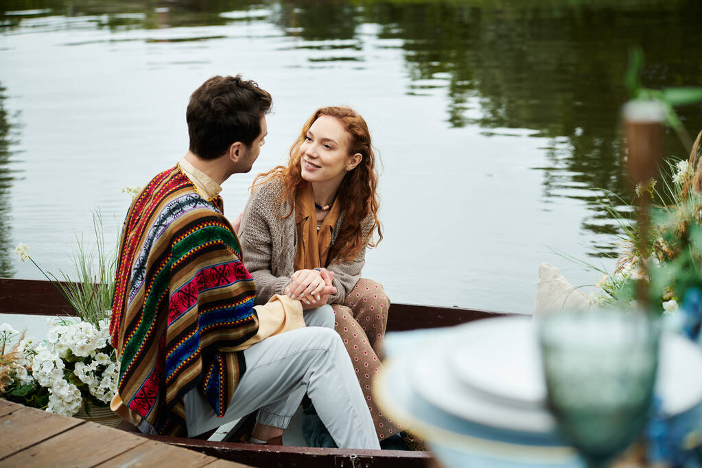 Un hombre y una mujer en traje boho se sientan pacíficamente en un barco en un lago tranquilo en un exuberante entorno de parque verde. - Foto, imagen