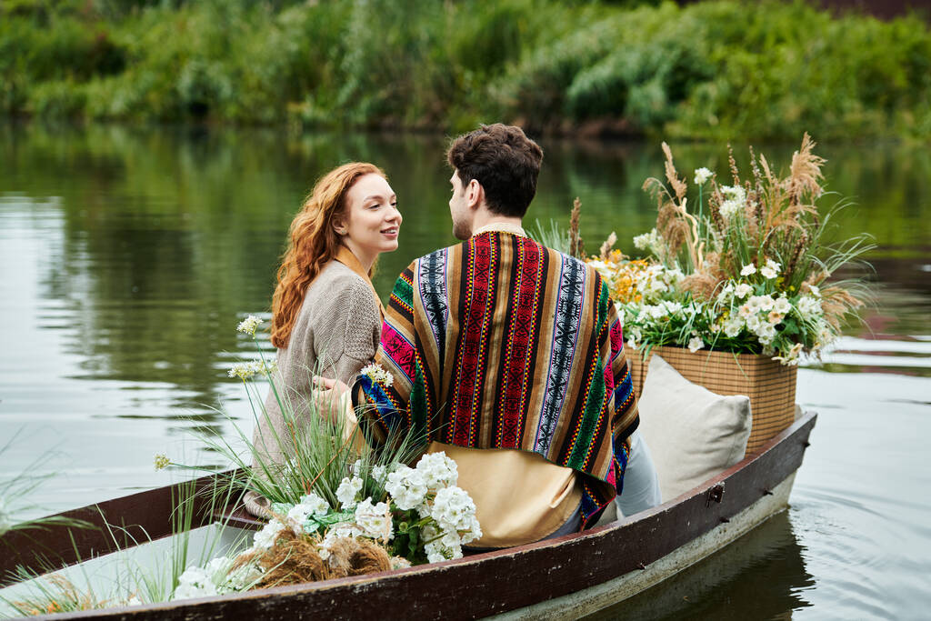 Ένας άντρας και μια γυναίκα στο Boho ντύνονται πλέουν σε μια βάρκα που περιβάλλεται από ανθισμένα λουλούδια, απολαμβάνοντας μια ρομαντική ημερομηνία σε ένα πράσινο πάρκο. - Φωτογραφία, εικόνα