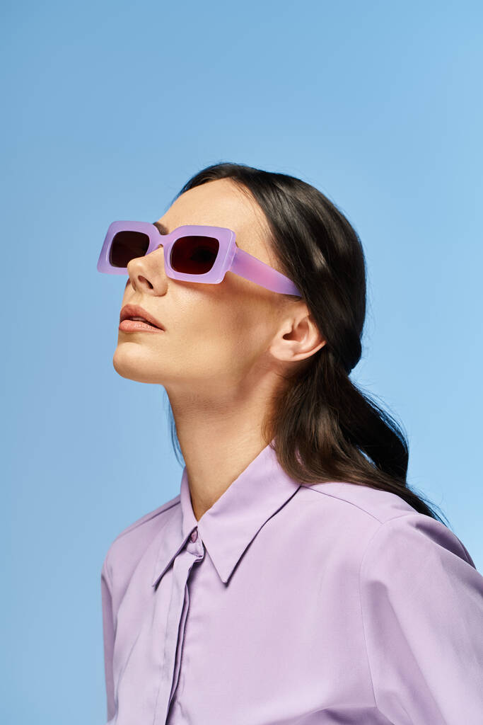 Una mujer de moda con una camisa púrpura y gafas de sol posa con confianza en un estudio sobre un fondo azul. - Foto, imagen