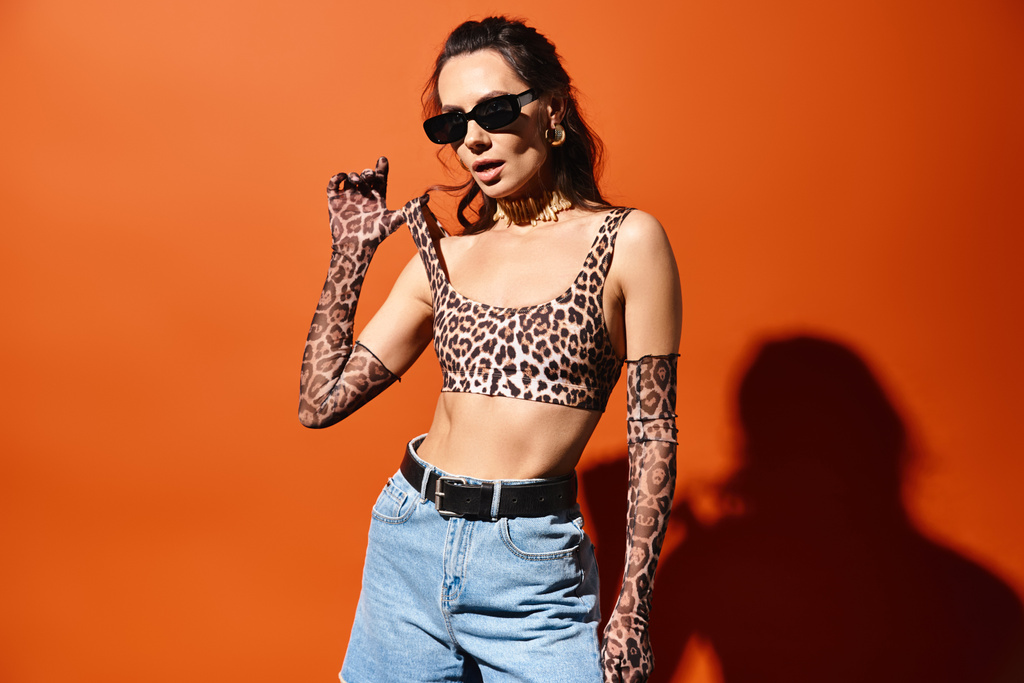 Mujer con estilo en gafas de sol posando con confianza en un top estampado de leopardo y jeans sobre un fondo naranja. - Foto, Imagen