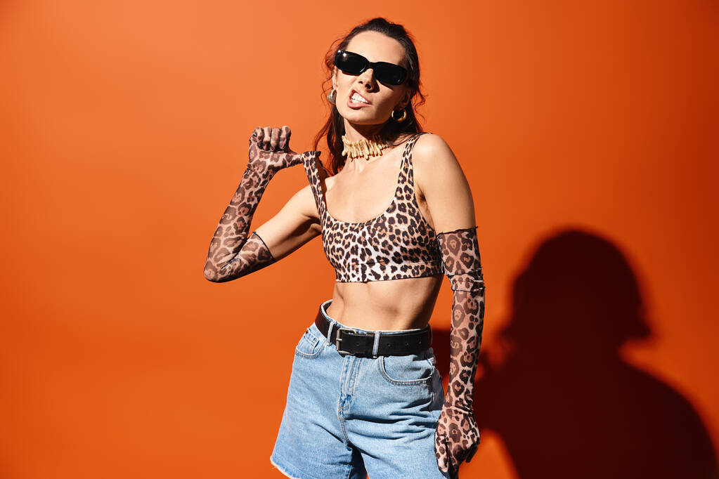 Tyylikäs nainen aurinkolaseissa seisoo luottavaisesti studiossa, yllään leopardi tulostustoppi ja denim-shortsit oranssia taustaa vasten. - Valokuva, kuva