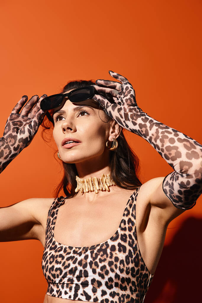 Uma mulher elegante em um top de impressão de leopardo segura as mãos em sua cabeça em um estúdio laranja vibrante durante uma sessão de moda de verão. - Foto, Imagem