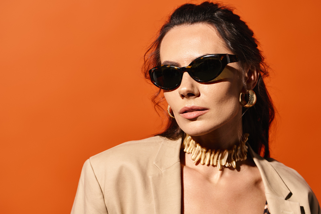 A stylish woman is seen wearing sunglasses and a tan jacket against an orange background, embodying summertime fashion. - Photo, Image