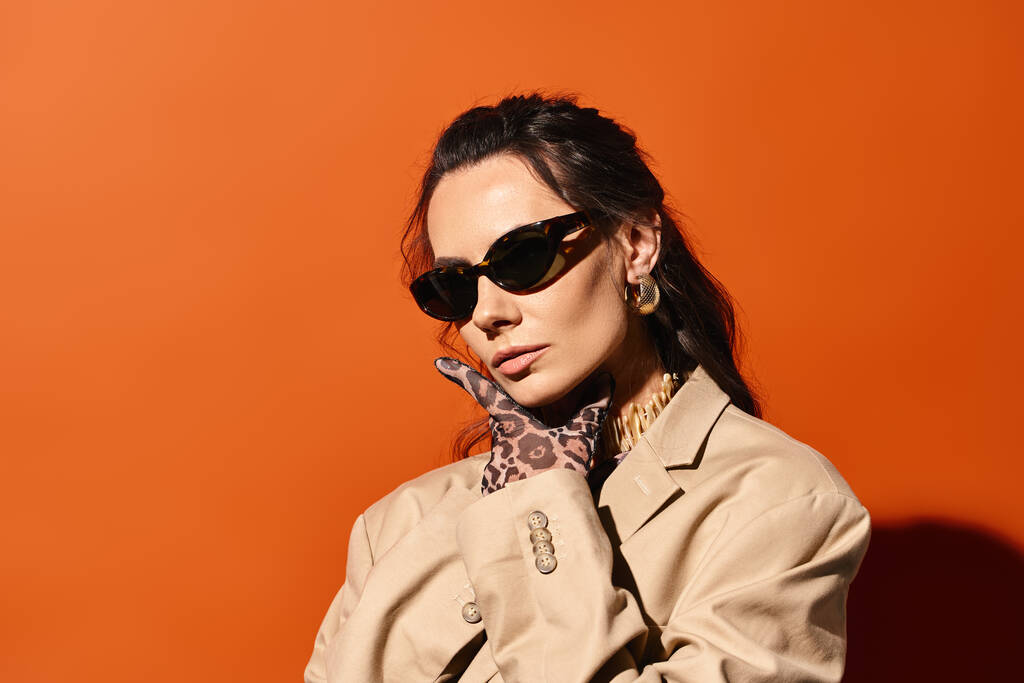 Uma mulher elegante em óculos de sol e um casaco de trincheira posa confiantemente contra um cenário de estúdio laranja vibrante, exalando chique de verão. - Foto, Imagem