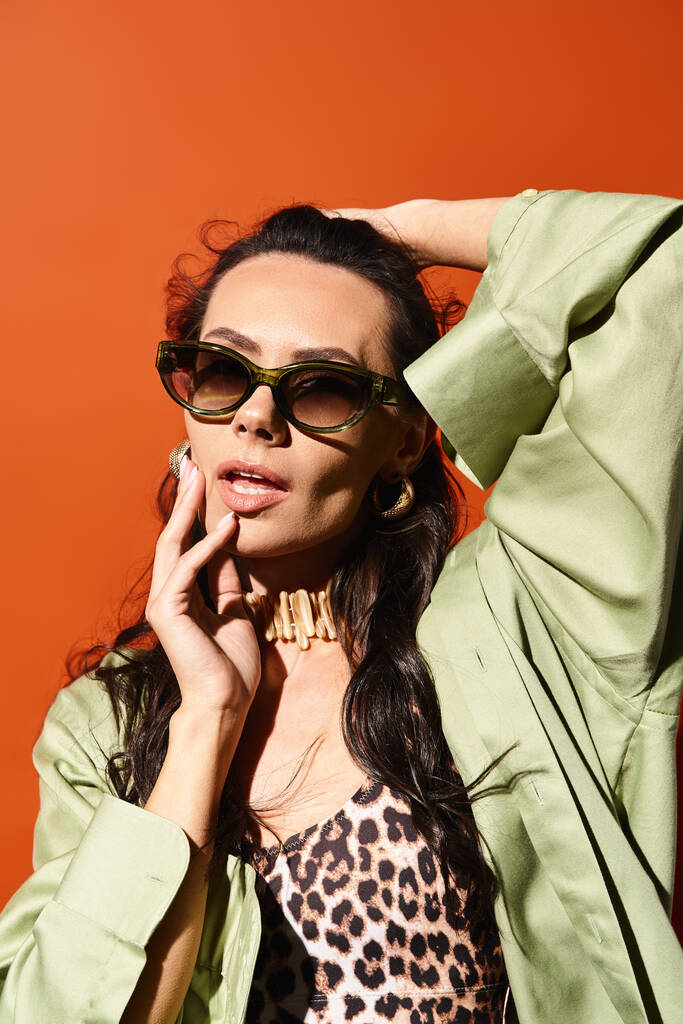 Egy elegáns nő napszemüveget és zöld dzsekit visel, narancssárga háttérrel pózol a stúdióban, nyári divatot árasztva.. - Fotó, kép