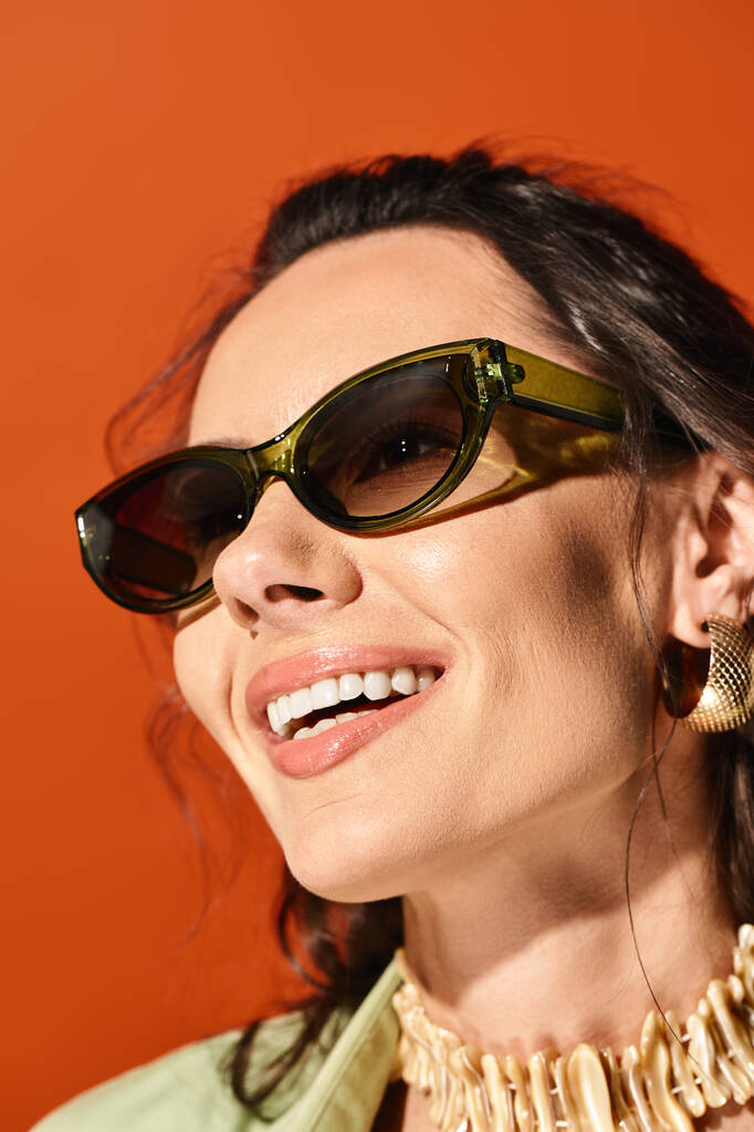 Una mujer de moda exuda elegancia veraniega en gafas de sol y un collar de declaración contra un fondo de estudio naranja. - Foto, imagen