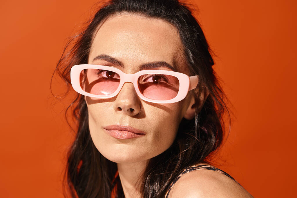 Μια όμορφη γυναίκα φοράει ροζ γυαλιά ηλίου και μια μαύρη μπλούζα ποζάρει σε ένα στούντιο σε πορτοκαλί φόντο, επιδεικνύοντας καλοκαιρινή μόδα.. - Φωτογραφία, εικόνα
