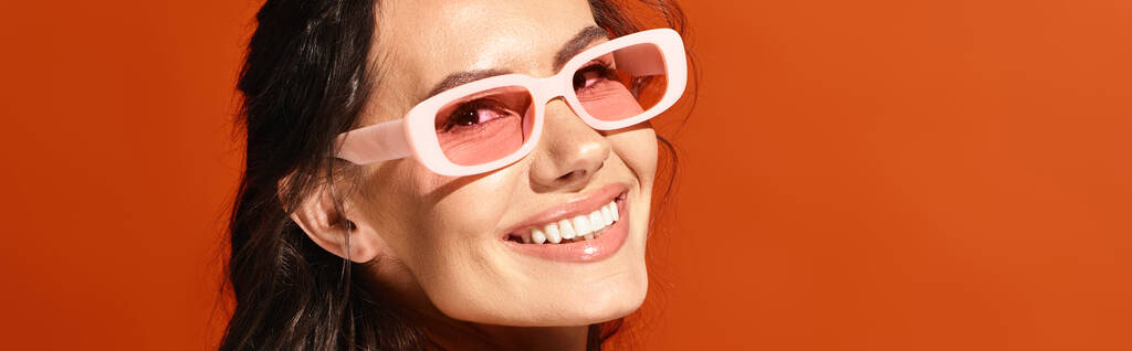 Una mujer elegante vestida con gafas de sol rosadas sonríe brillantemente a la cámara en un vibrante entorno de estudio sobre un fondo naranja. - Foto, Imagen