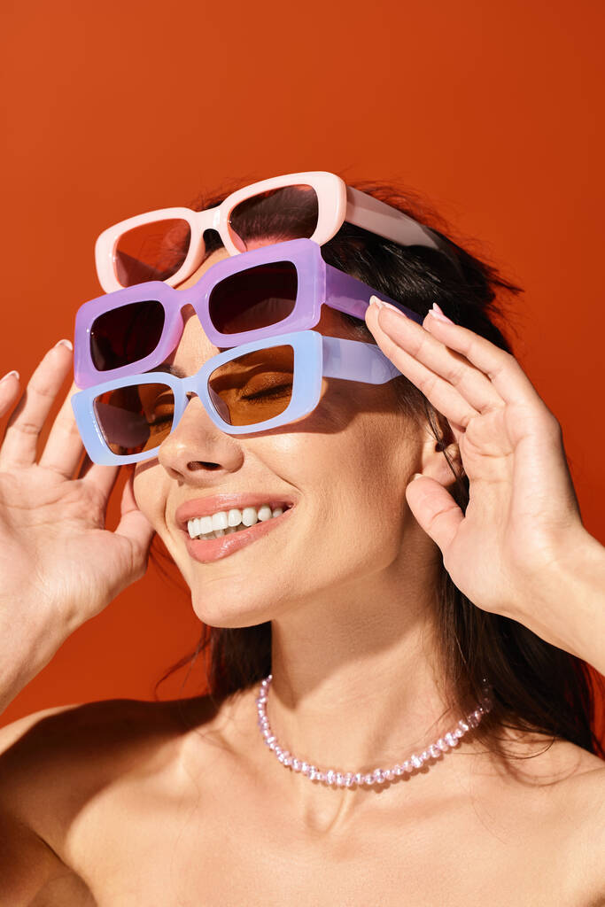 Une femme à la mode portant des lunettes de soleil et un collier, respirant la confiance dans un studio lumineux sur fond orange. - Photo, image