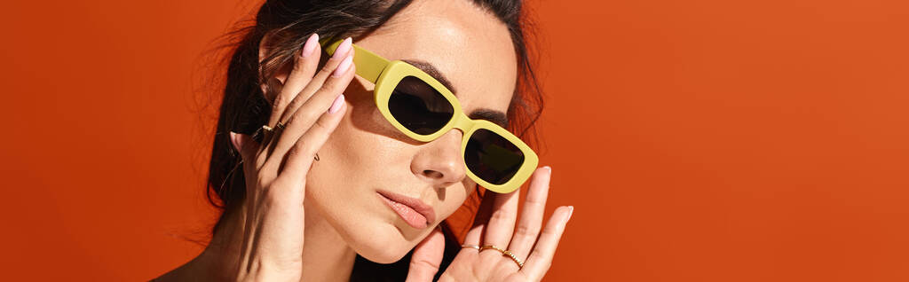 Una donna elegante che indossa occhiali da sole gialli posa con le mani delicatamente posizionate sul suo viso, trasudando fiducia e moda estiva su uno sfondo arancione studio. - Foto, immagini