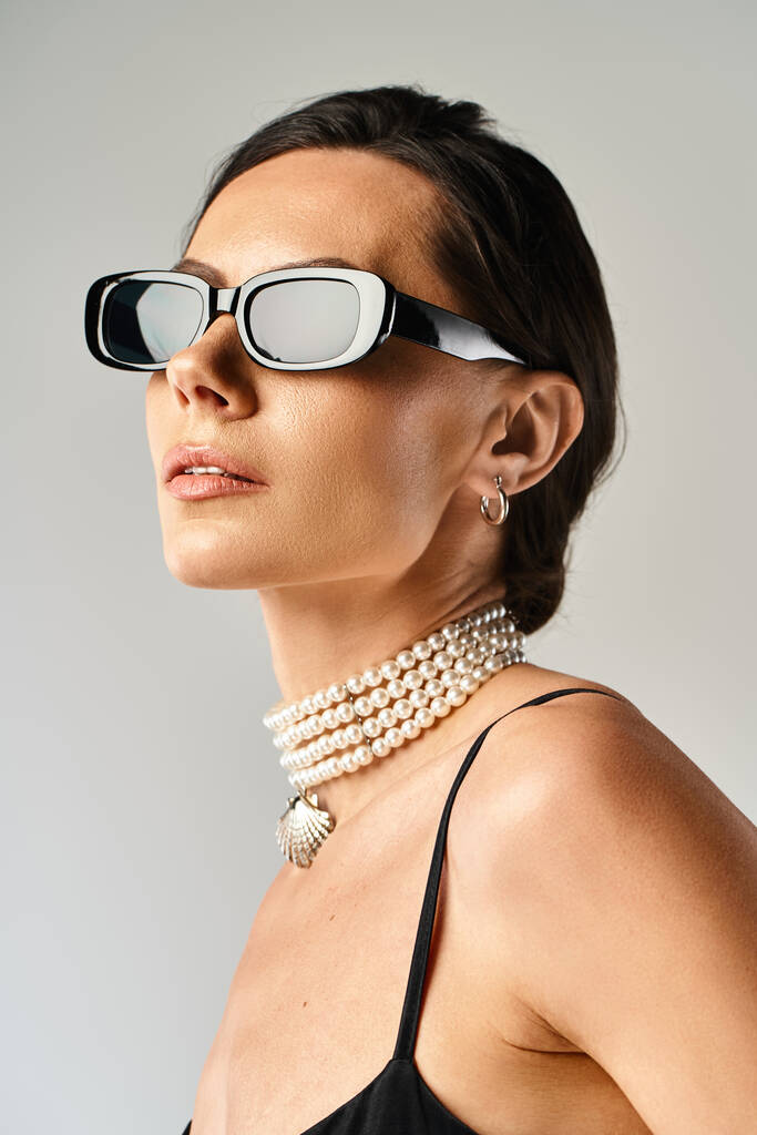 Eine stilvolle Frau ziert eine Sonnenbrille und Perlen und strahlt Eleganz und Raffinesse vor grauem Hintergrund aus. - Foto, Bild