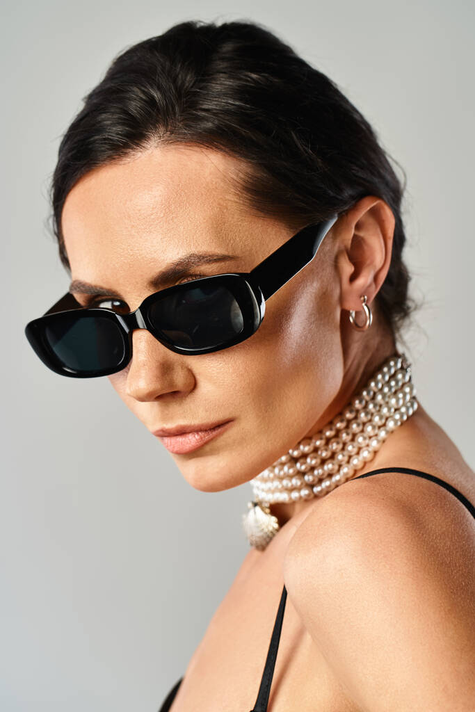 Модна жінка з перлами навколо шиї вражає позу в сонцезахисних окулярах на сірому фоні. - Фото, зображення