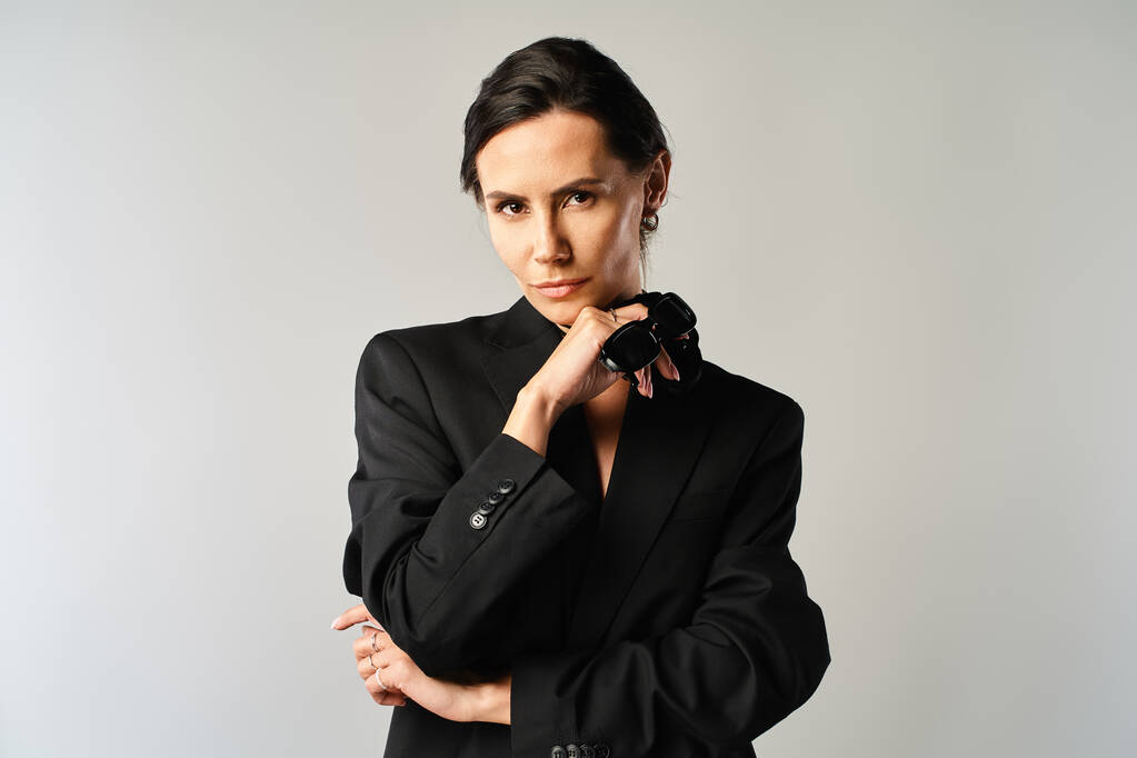 A fashionable woman in sunglasses confidently poses in a sleek black suit against a grey studio backdrop. - Photo, Image