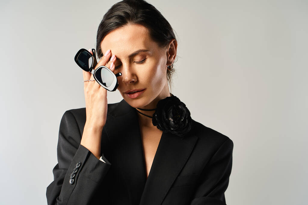 Uma mulher na moda em um terno preto segura com confiança um par de óculos contra um pano de fundo cinza. - Foto, Imagem