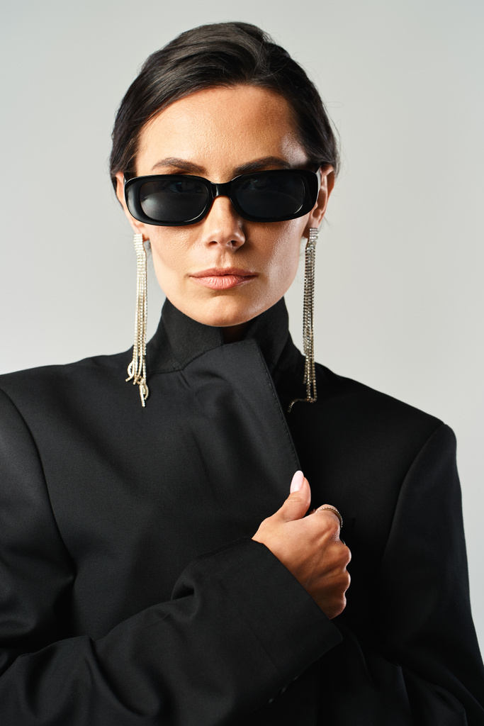 Une femme élégante avec des lunettes de soleil et une veste noire pose en toute confiance dans un studio sur un fond gris. - Photo, image