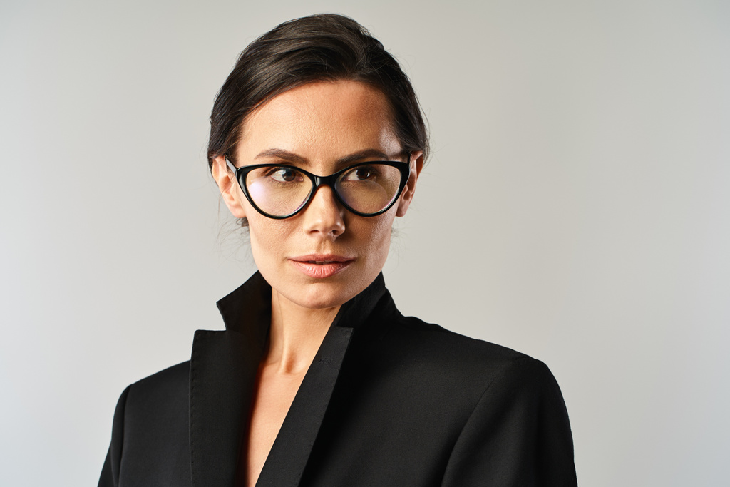 Egy elegáns nő kifinomultságot áraszt fekete kabátban és szemüvegben, szürke stúdióháttérrel.. - Fotó, kép