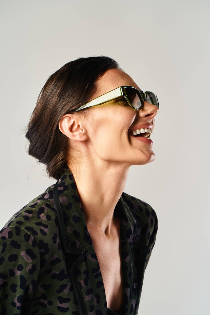 Uma mulher elegante exibe com confiança uma camisa de impressão de leopardo e óculos de sol na moda em um cenário de estúdio contra um fundo cinza. - Foto, Imagem