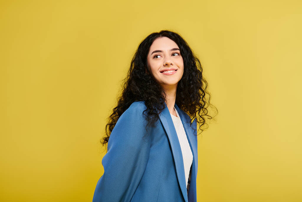 青いジャケットの若いブルネットの女性は,スタイルと感情を除く印象的な黄色い壁の前に自信を持って立っています.. - 写真・画像