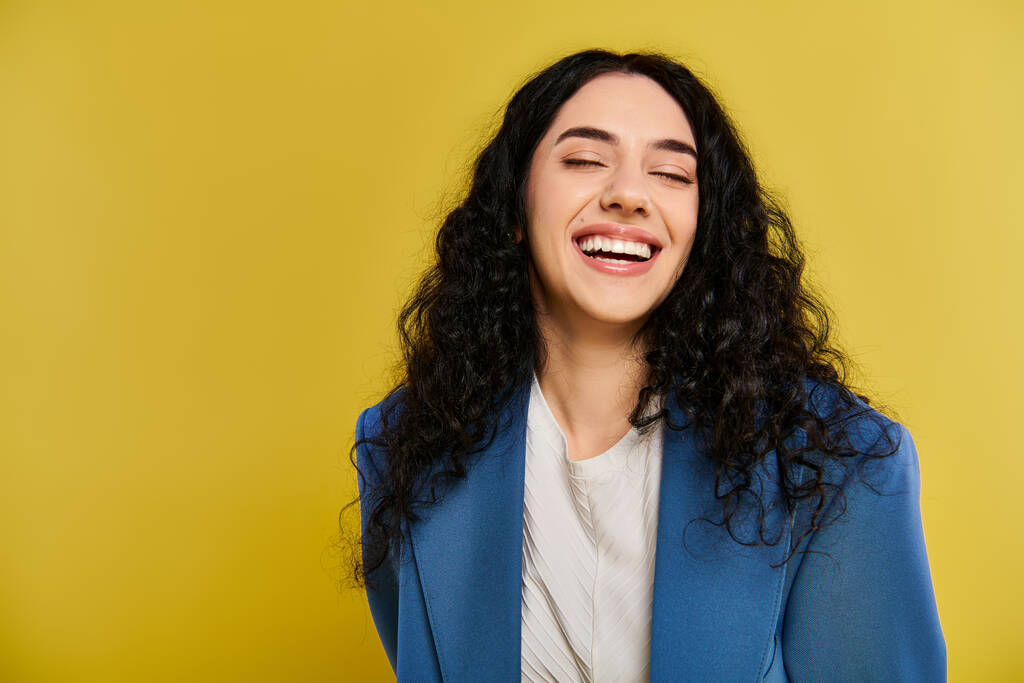 Een jonge vrouw met krullend haar glimlacht en draagt een blauw jasje, straalt vreugde en vertrouwen uit in een studio met een gele achtergrond. - Foto, afbeelding