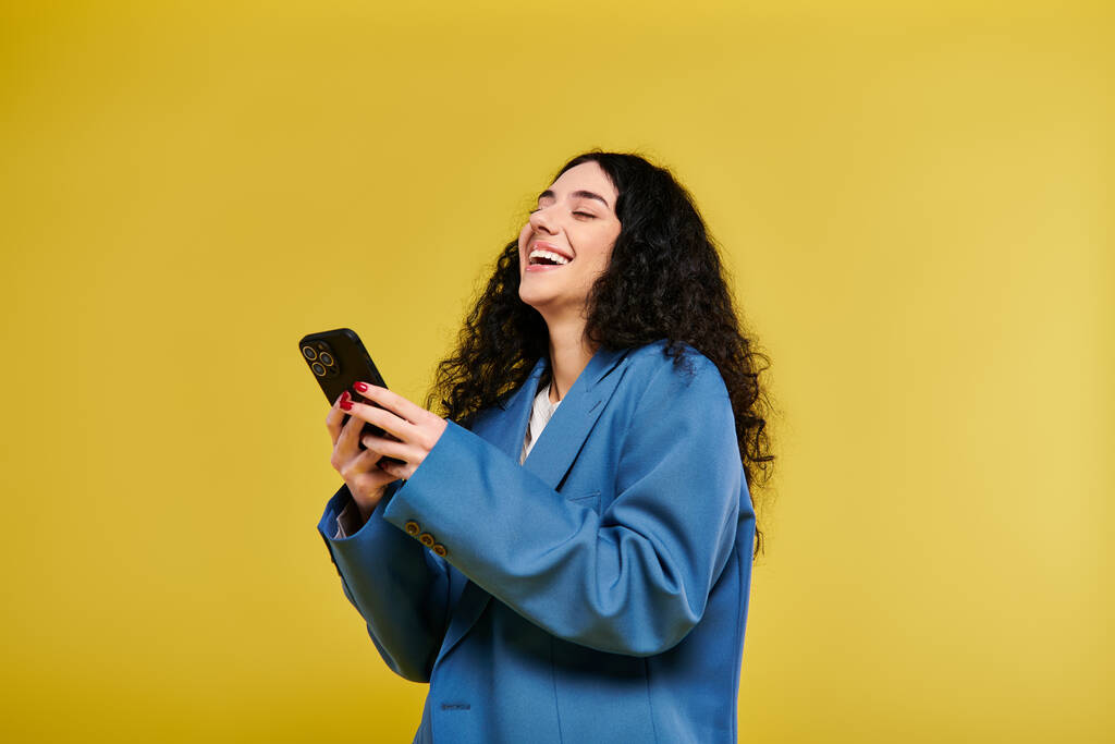 Egy elegáns, göndör hajú, kék kabátos fiatal nő mobiltelefont tart fényes sárga háttér előtt.. - Fotó, kép