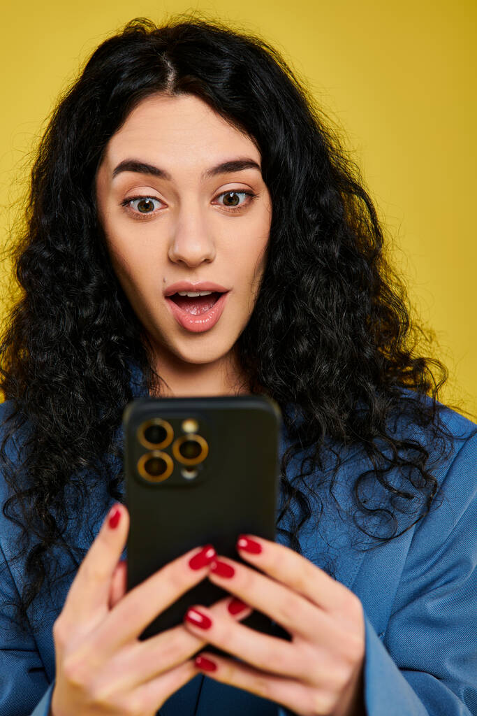 Eine junge brünette Frau mit lockigem Haar, stilvoll gekleidet, hält vor gelbem Hintergrund ein Mobiltelefon mit überraschendem Gesichtsausdruck. - Foto, Bild