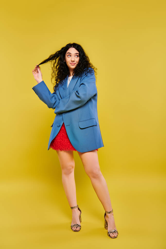 Una donna bruna con i capelli ricci in posa in una giacca blu e gonna rossa, trasudante stile e fascino su uno sfondo giallo vibrante. - Foto, immagini