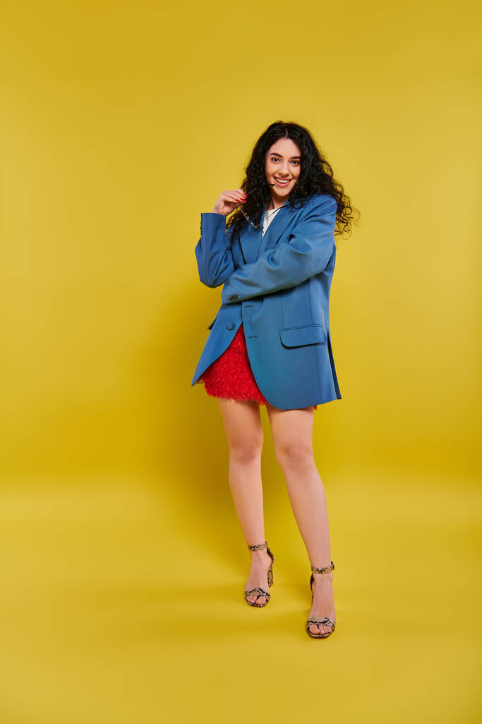 Een jonge brunette vrouw met krullend haar poseert vol vertrouwen in een stijlvolle blauwe jas en rode rok, uitstralen gratie en charme tegen een levendige gele achtergrond. - Foto, afbeelding